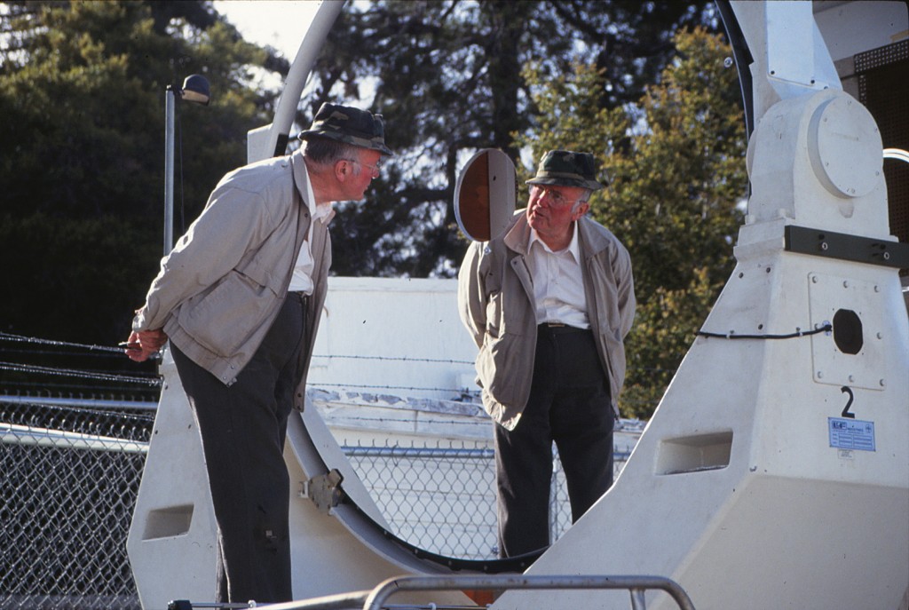 A man looking into a large round telescope mirror and seeing his own reflection.